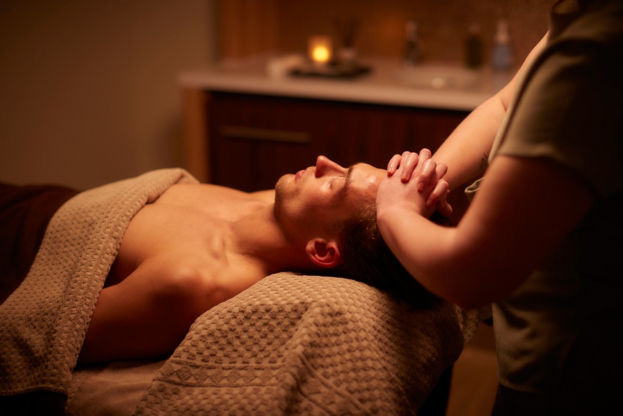 Person lies on a massage table, receiving a forehead and scalp massage, draped in a textured towel within a dim, warm spa room featuring a candle and small bottles.